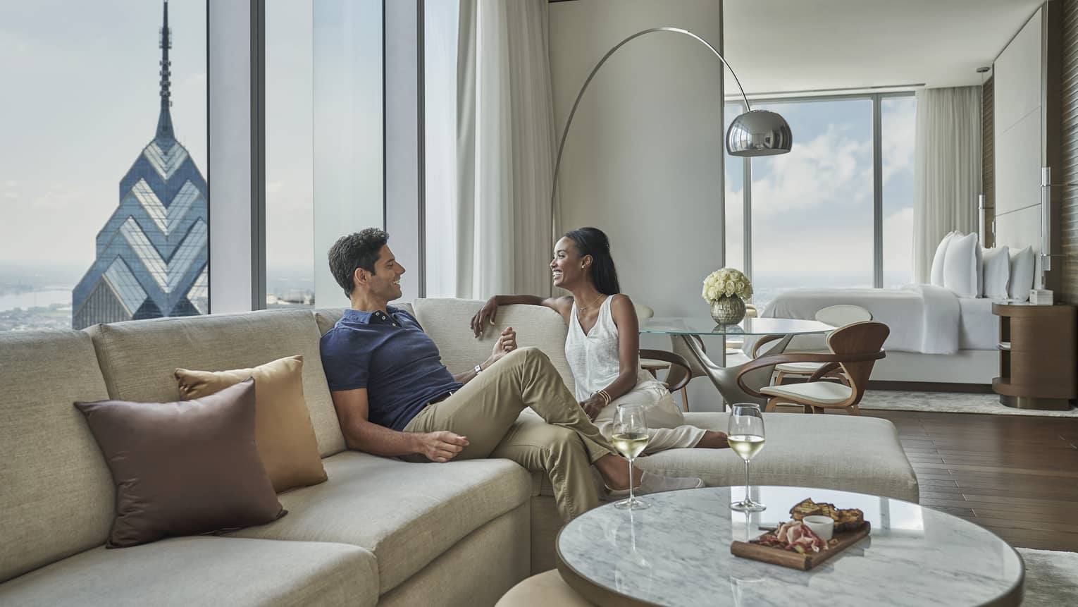 A man and woman lounge on a tan sectional in their modern, light and airy living room adjacent to their bedroom 