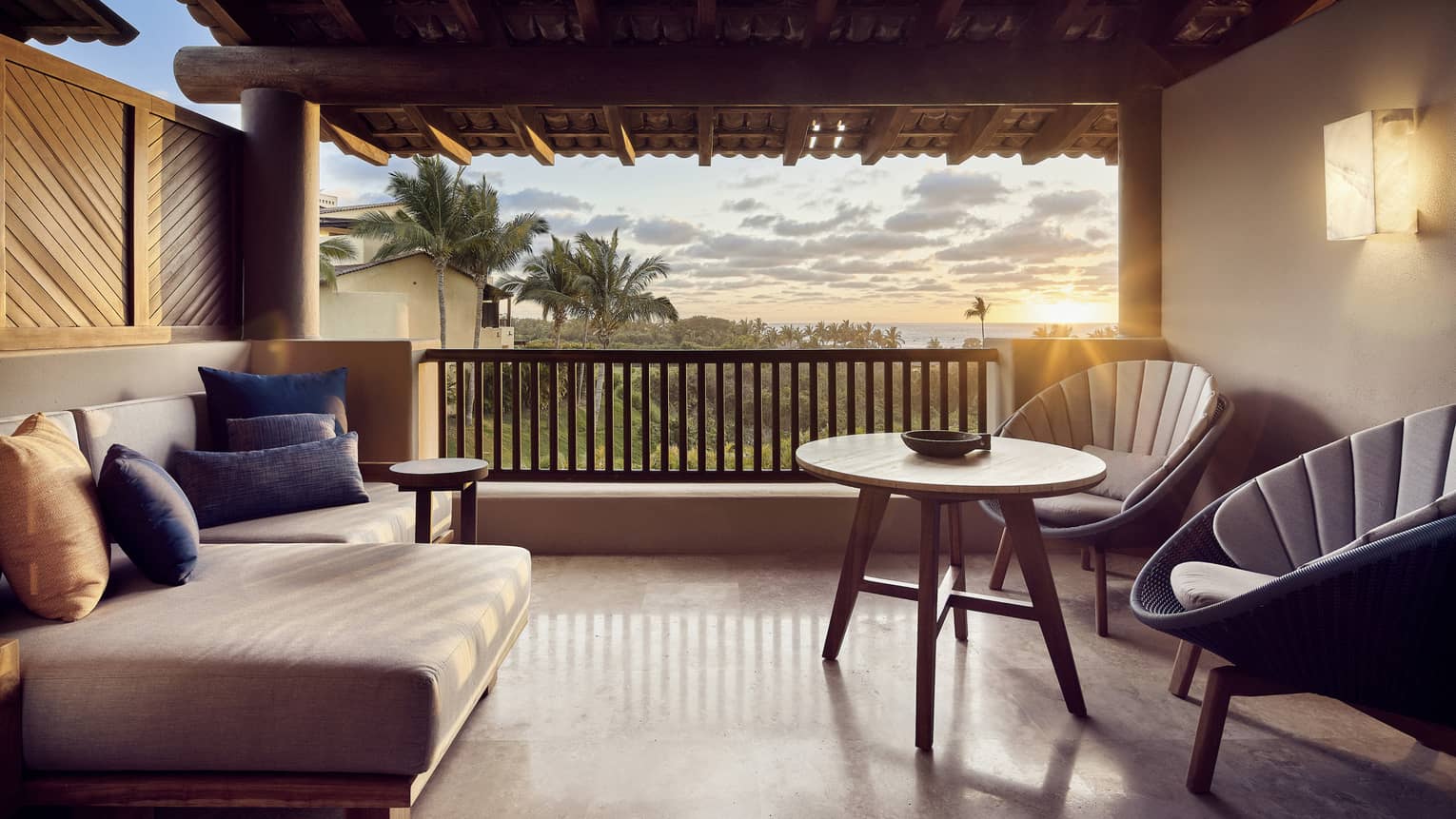 A sunlit hotel balcony with two chairs, a small table, and a sofa.