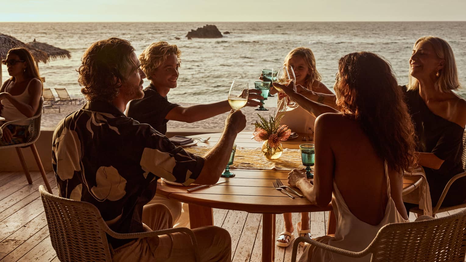 Five guests raise their glasses in a toast around a round wood table on a wood deck overlooking the beach and ocean.