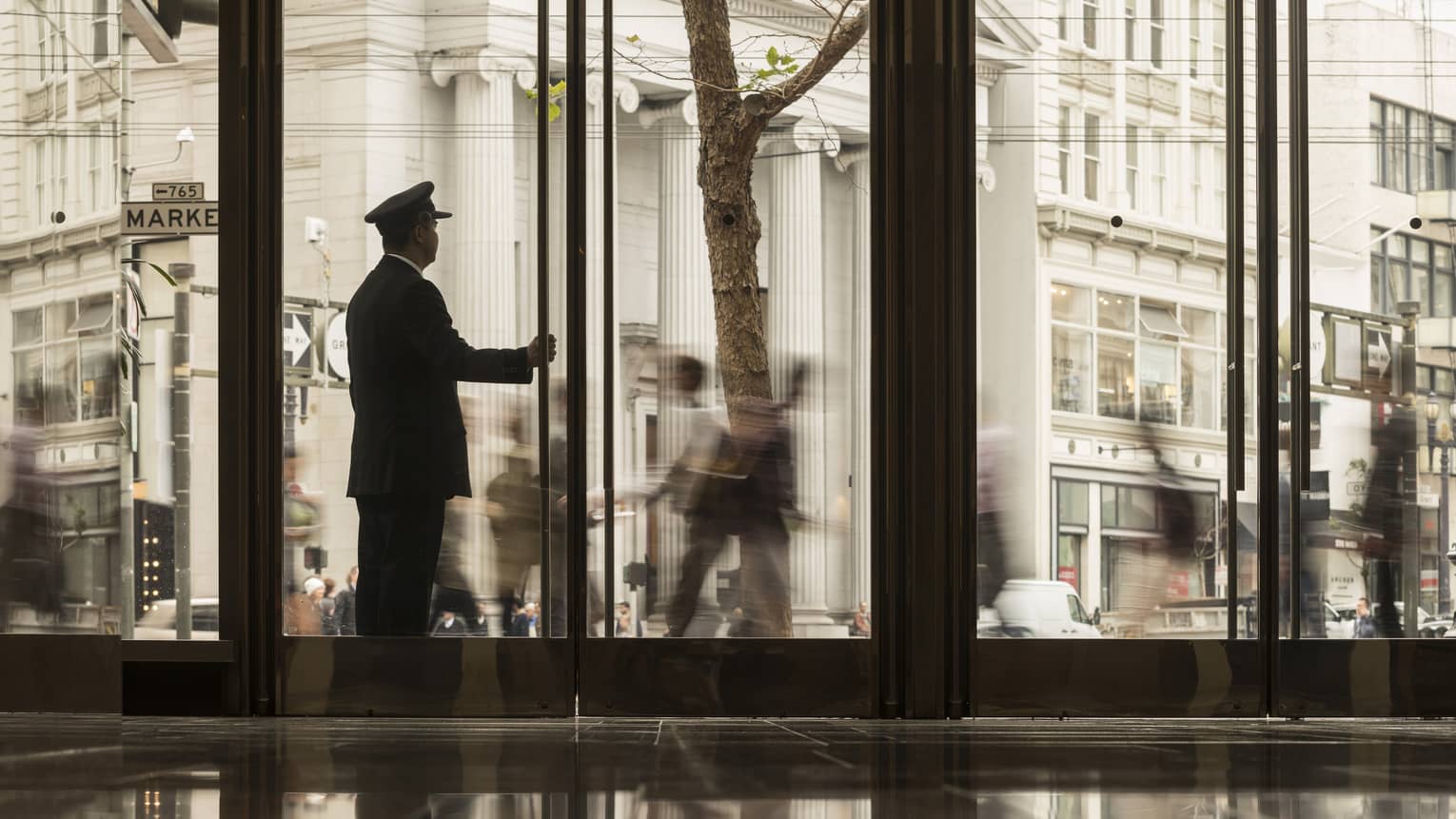 A doorman holding the door of a hotel lobby while looking outside at people walking around the city.