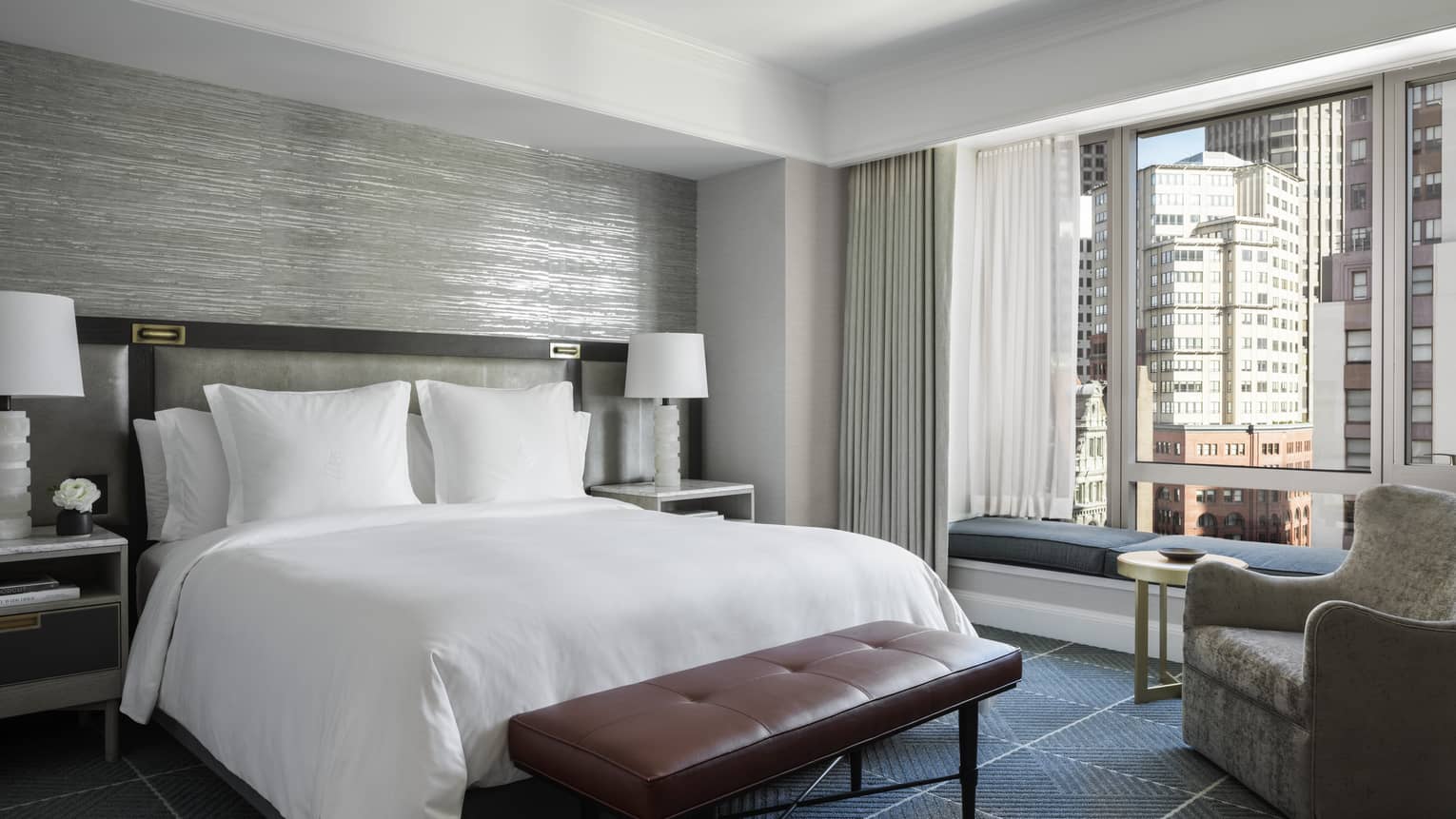 A hotel room with a bed with white sheets, a red bench, a grey chair, two white lamps, and a large window looking out at a city.