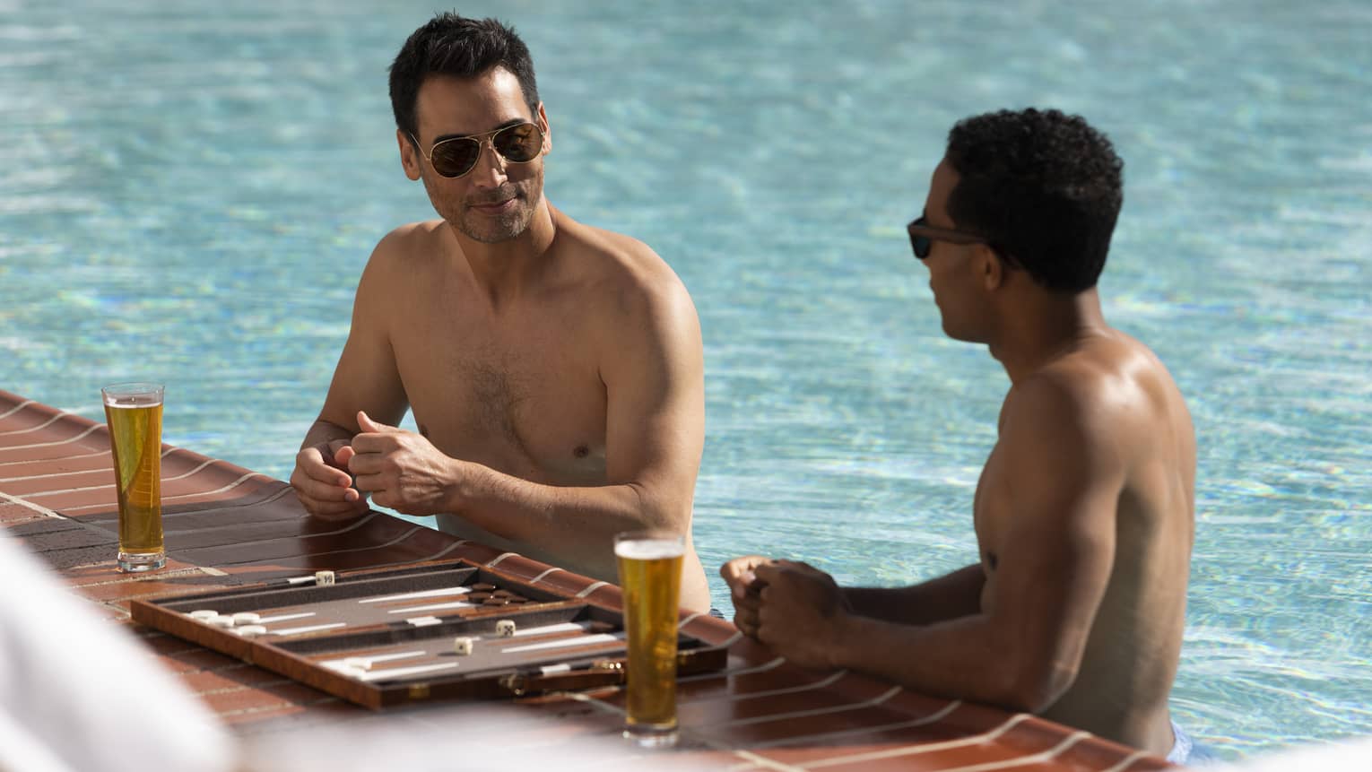 Two people wearing sunglasses and standing in a swimming pool, right at the edge, with clear drink glasses containing a golden liquid and a game board
