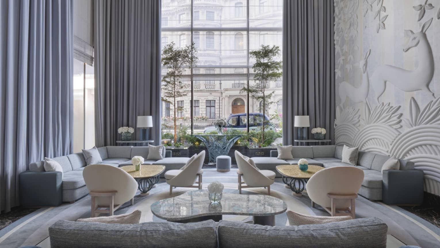A luxurious and elegantly decorated lobby at the Four Seasons Hotel in London's Mayfair district. The room features two large blue velvet sofas forming a U-shape around a modern coffee table, flanked by beige armchairs. Intricate wall art and large floor-to-ceiling windows overlooking a classic London street scene enhance the sophisticated atmosphere. The decor includes soft grey curtains, patterned rugs, and tasteful lighting fixtures, creating a serene and upscale ambiance.,A luxurious and elegantly decorated lobby at Four Seasons Hotel in London's Mayfair district. The room features two large blue velvet sofas, forming a U-shape around a modern coffee table, flanked by beige armchairs. Intricate wall art and large floor-to-ceiling windows overlooking a classic London street scene enhance the sophisticated atmosphere. The decor includes soft grey curtains, patterned rugs and tasteful lighting fixtures, creating a serene and upscale ambiance.