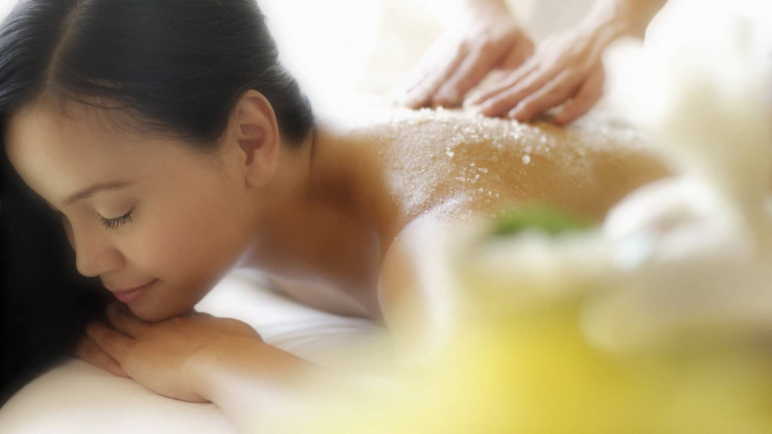 Woman lying down with eyes closed as hands massage salt scrub into shoulders