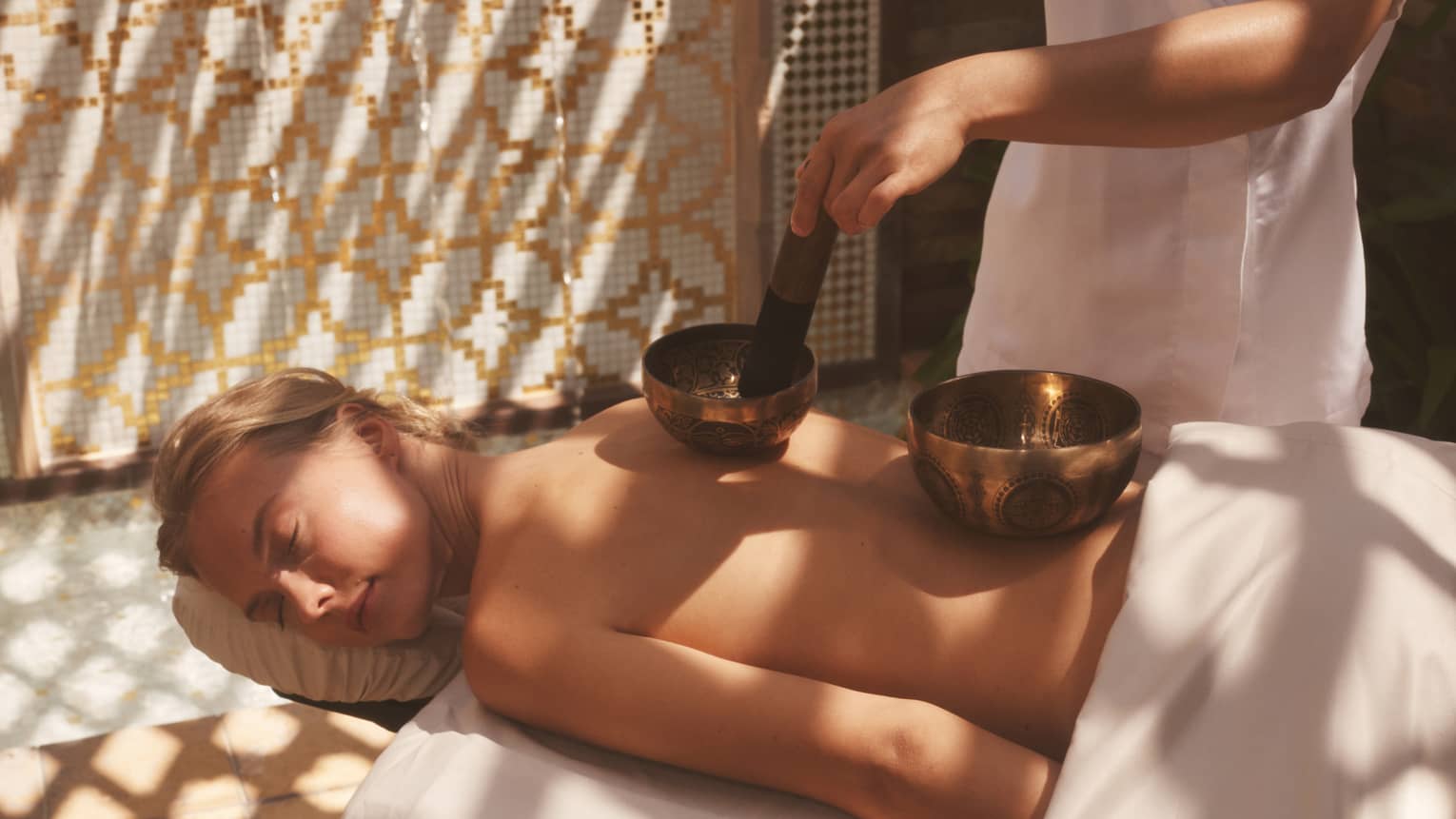 Unclothed person lying prone on a spa table with two sound healing bowls on back and spa therapist using them