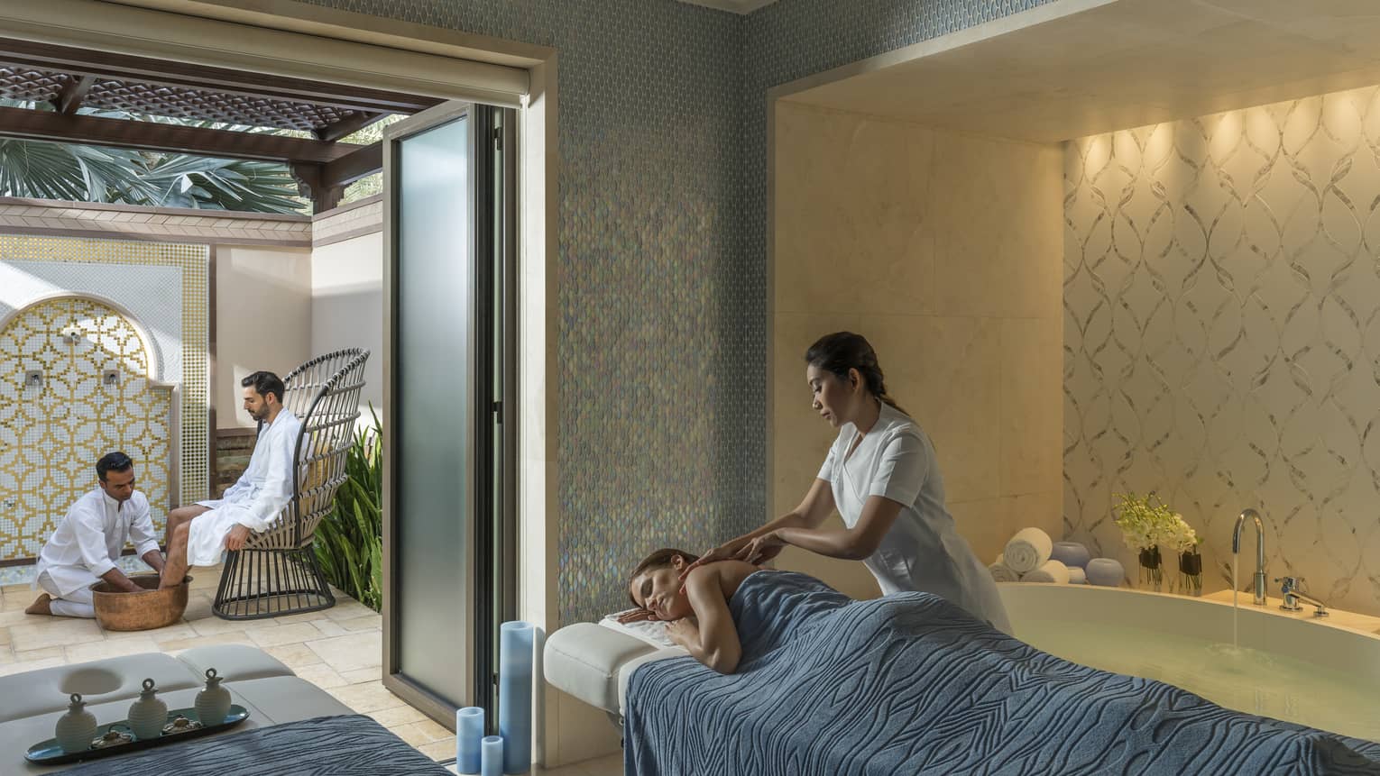 A woman under a blue sheet gets a massage from a masseuse surrounded by candles and natural light. 