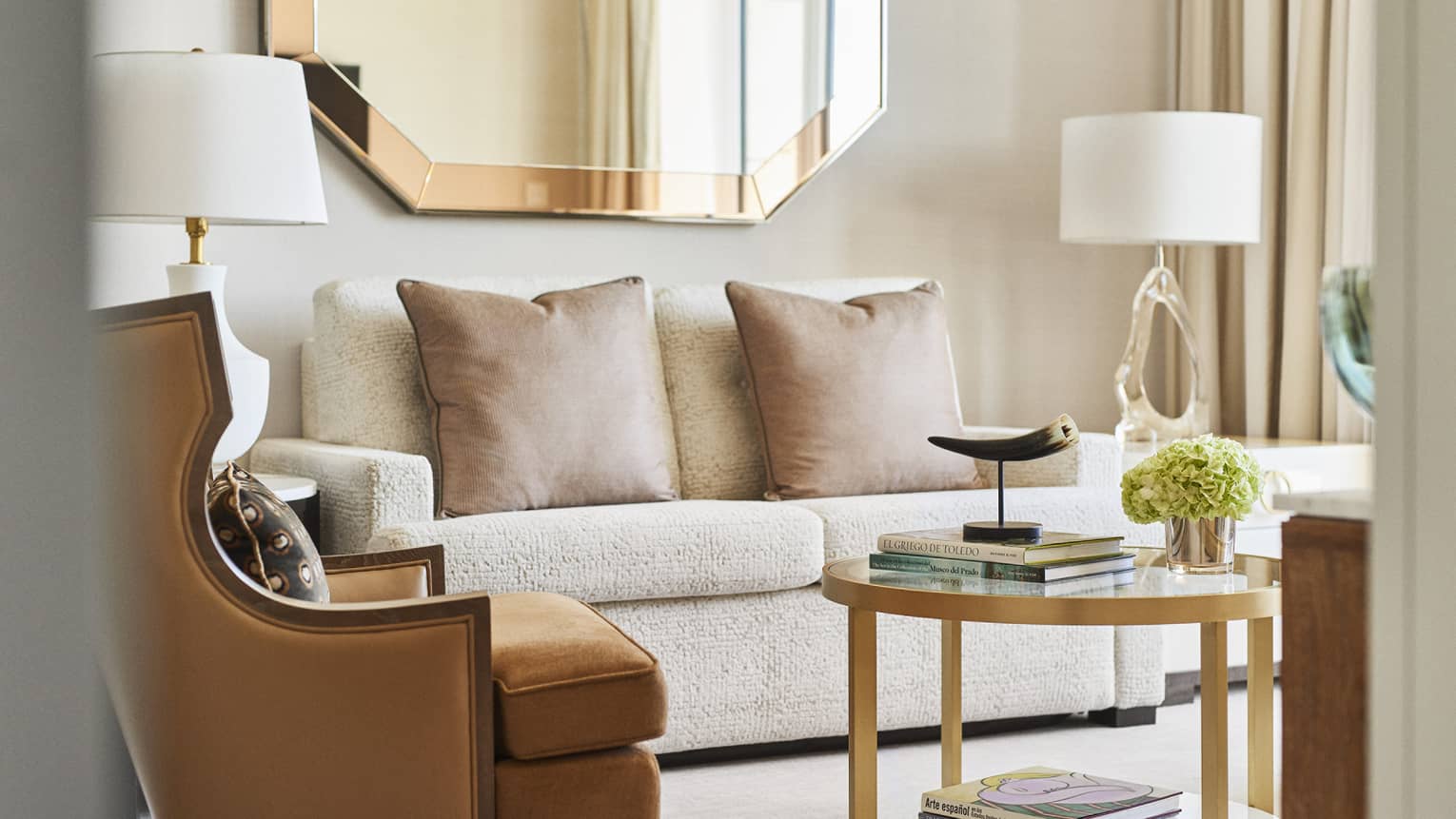 Close-up of a beige sofa with two cushions, brown armchair, gold-framed mirror and a round coffee table with books, horn sculpture and floral arrangement