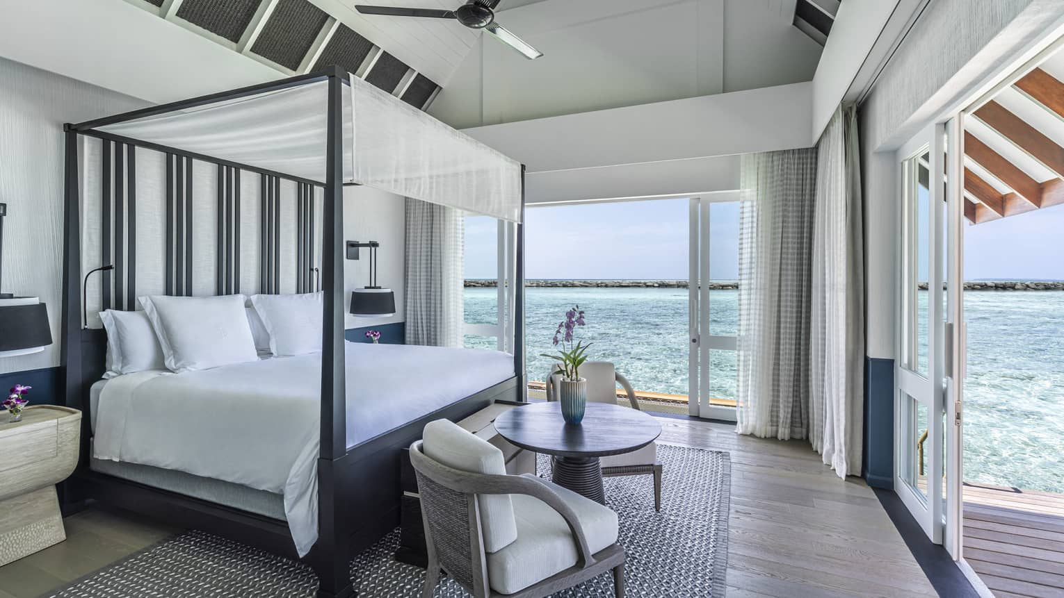 Bedroom with four-poster canopied bed, two-seater round table, double doorway leading to deck with ocean views