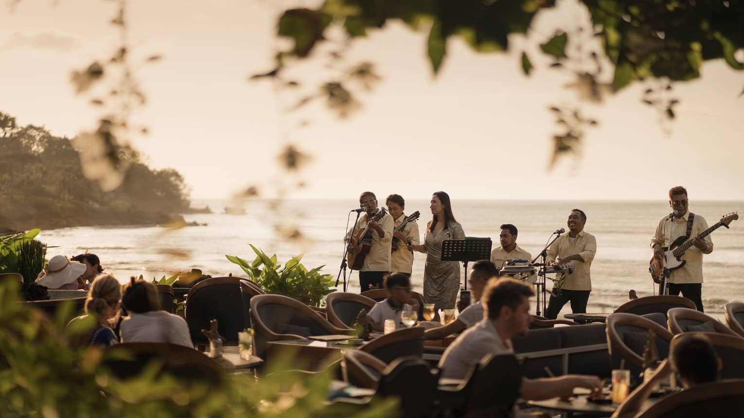 View through foliage of a musical band standing with their backs to the ocean and playing for dining guests at dusk.
