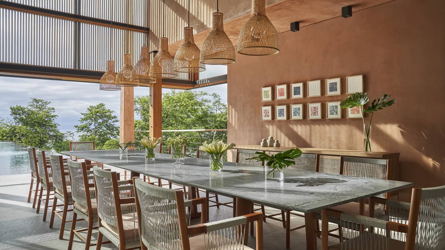 Long dining table with 12 chairs, whicker lighting fixture, terracatta-coloured wall lined with small square framed prints