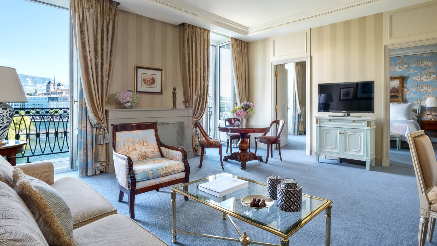 Spacious hotel suite with a beige sofa, glass coffee table, antique-style armchair, a round dining table, striped wallpaper, TV and a balcony with a lake view