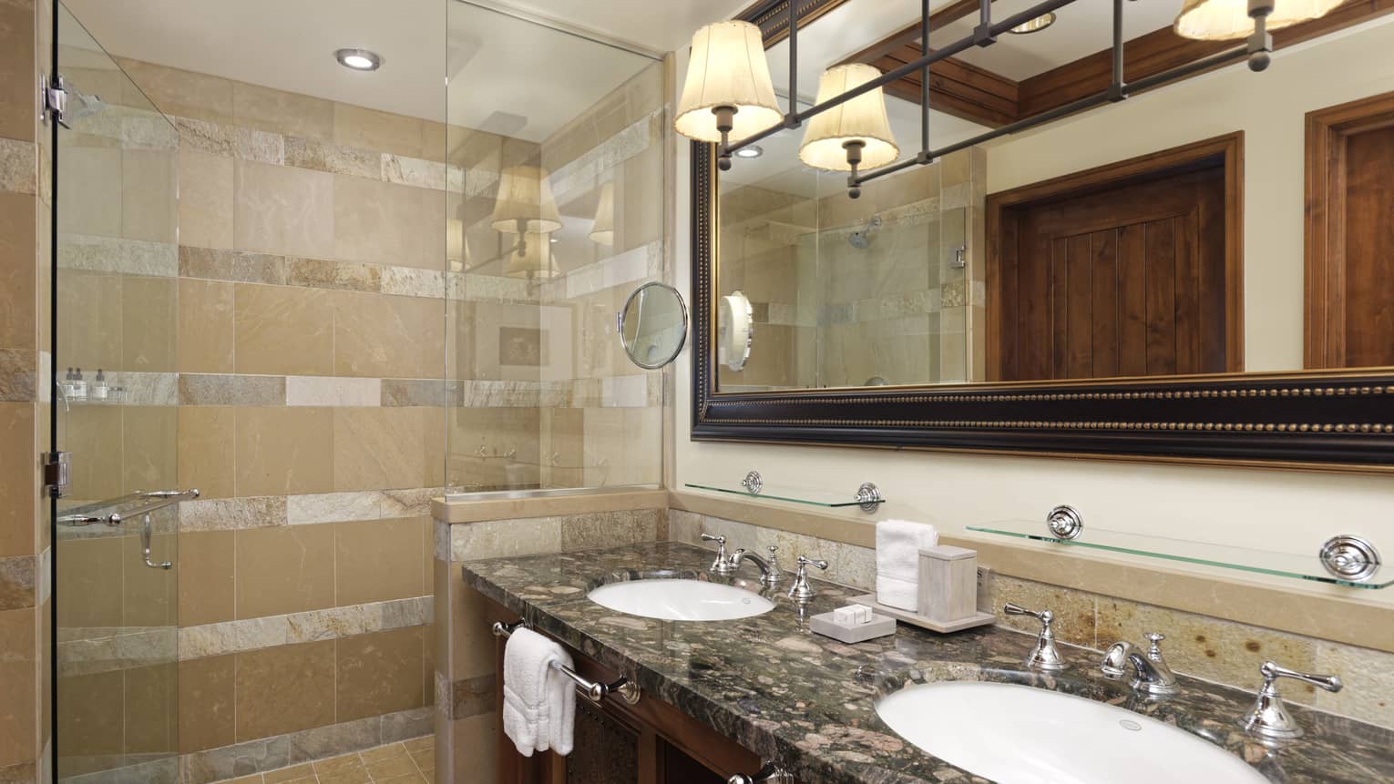 Luxury bathroom with a dark granite double vanity, large framed mirror, pendant lights and a glass-enclosed shower featuring beige and tan stone tiles
