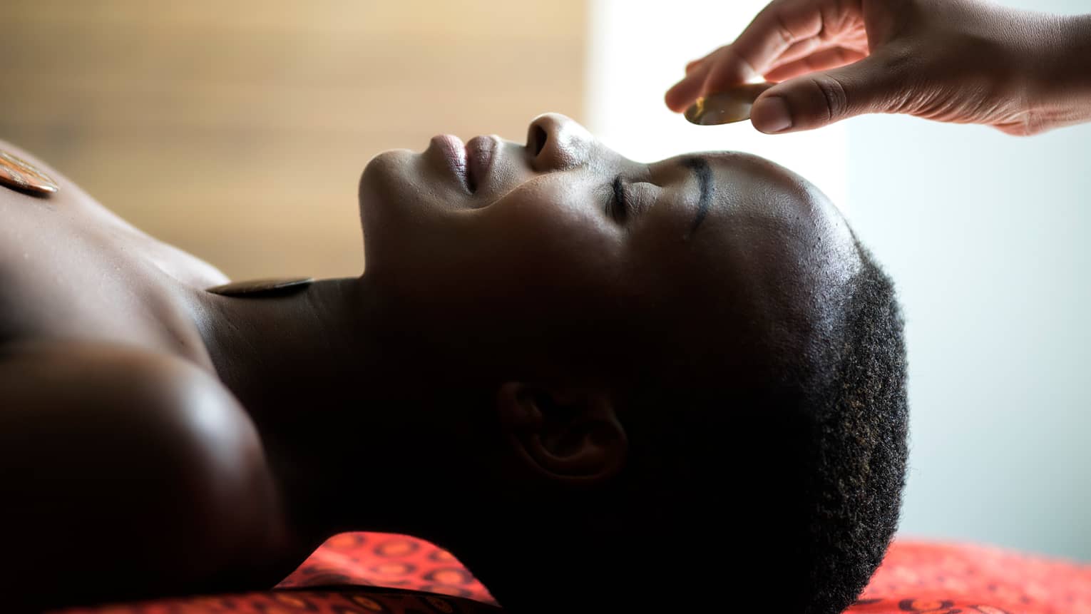 Hand placing stone on woman's forehead as she smiles, face-up on spa table