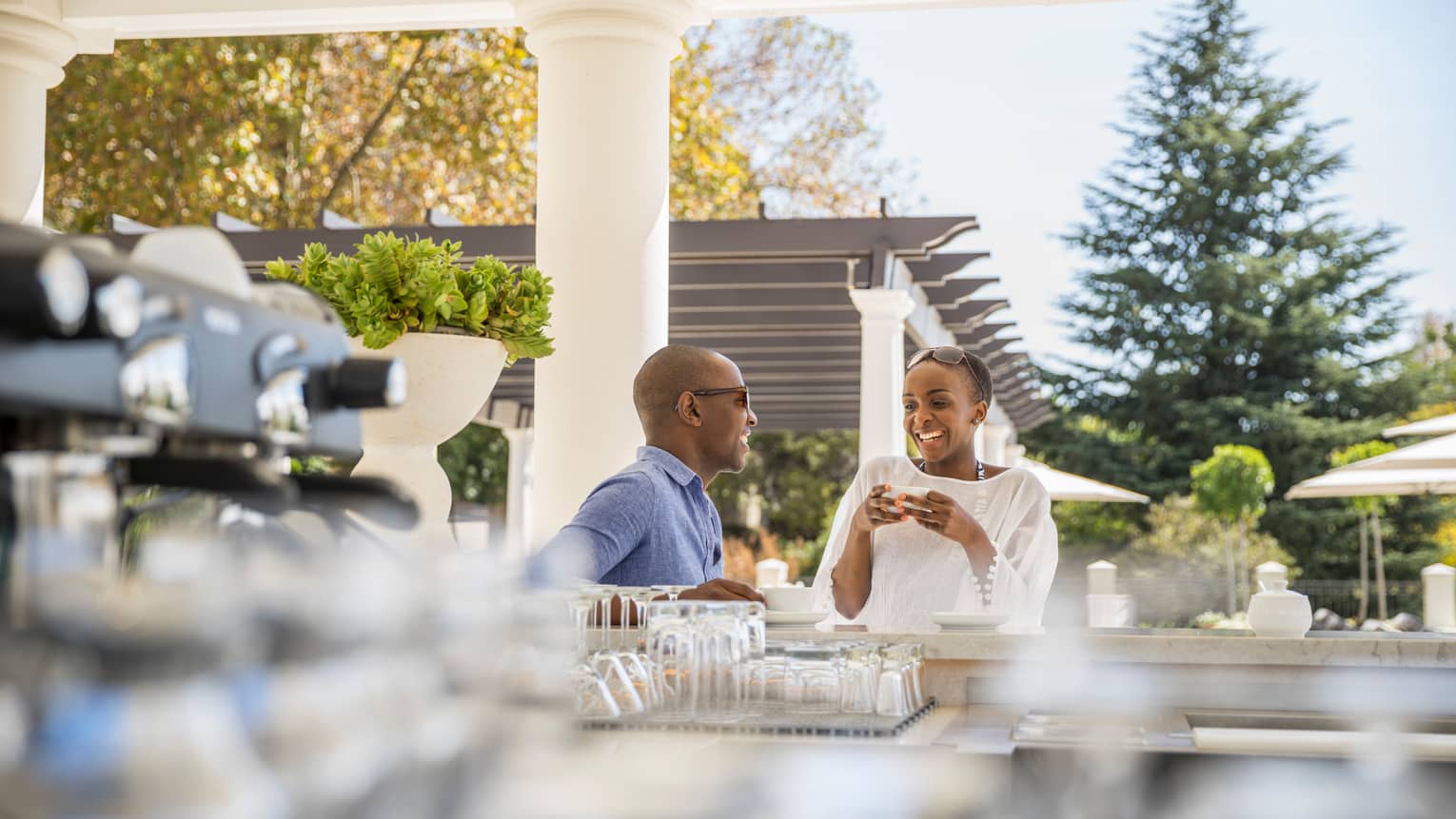 Man and woman drink espresso coffee at bar on sunny patio, white pergola in background