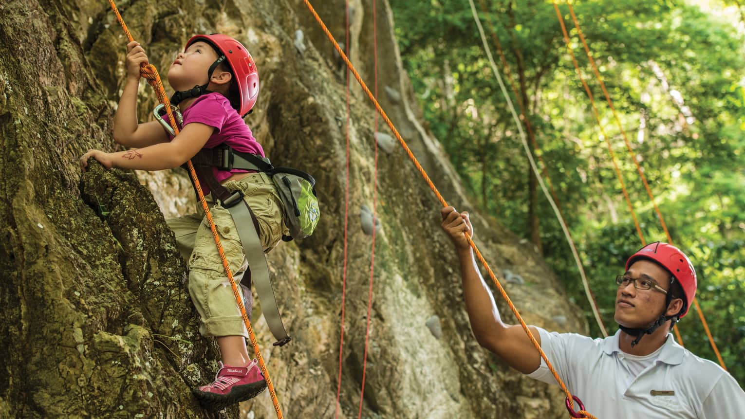 Man holds rope, helps young girl wearing helmet climb rock face