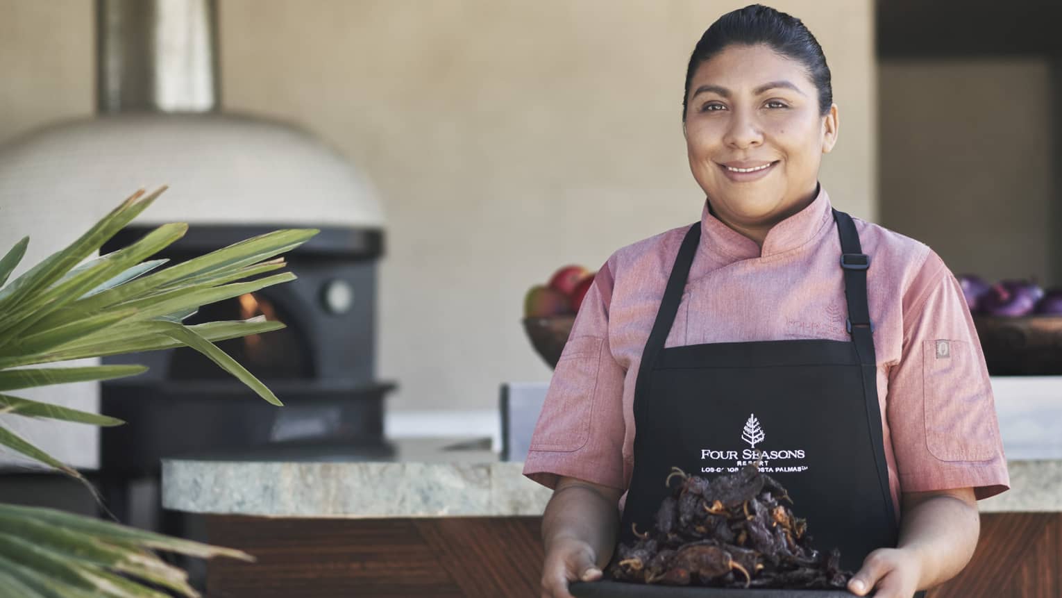 A chef in a pink shirt and black apron holding a plate of meat in front of an outdoor kitchen.