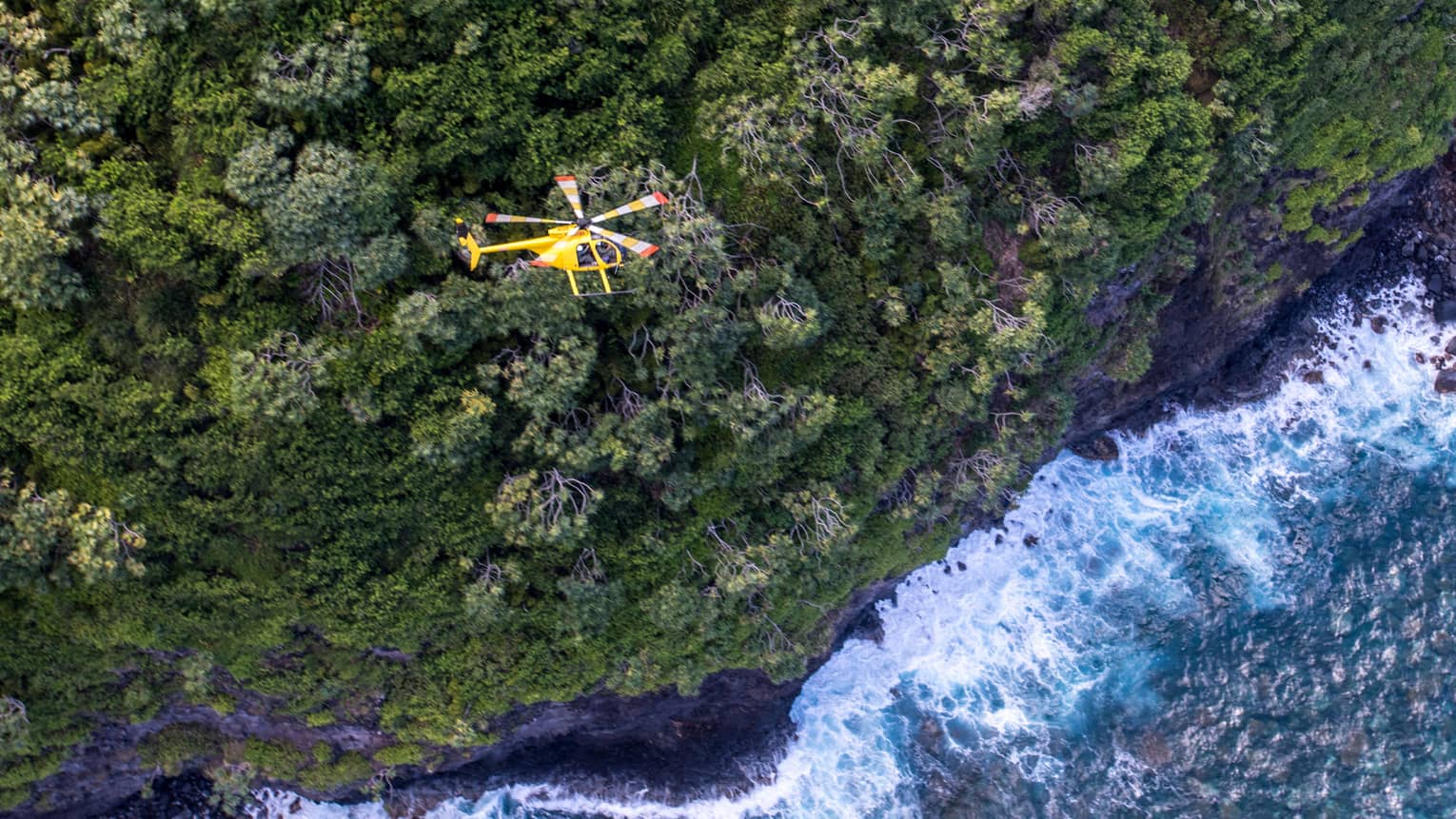 A yellow helicopter flies over a steep forested mountain. Powerful waves crash against the shoreline in the water below.