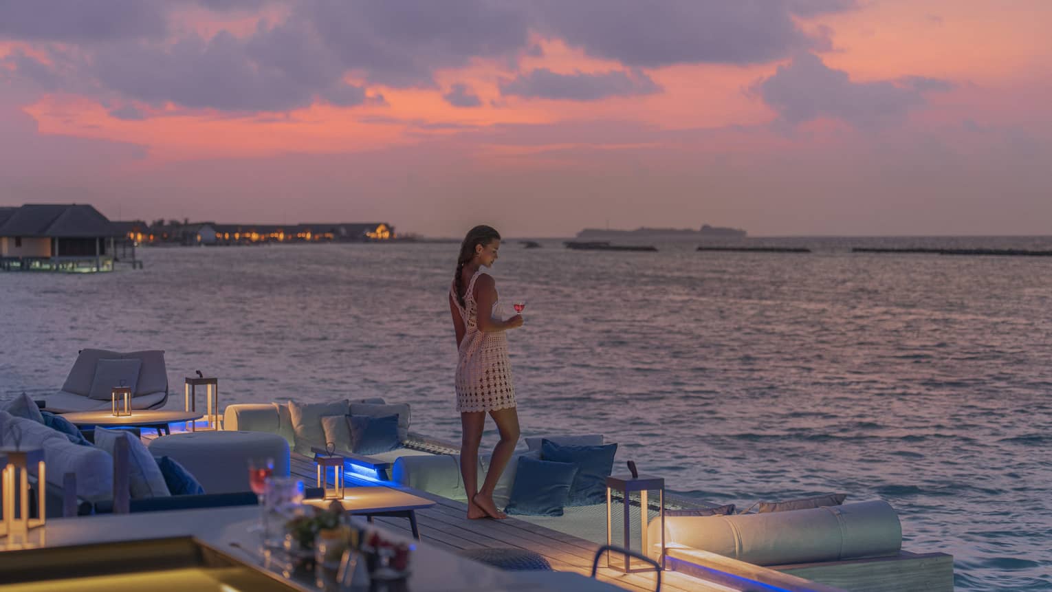 Woman holding cocktail stands at edge of Seabar overlooking the water at sunset