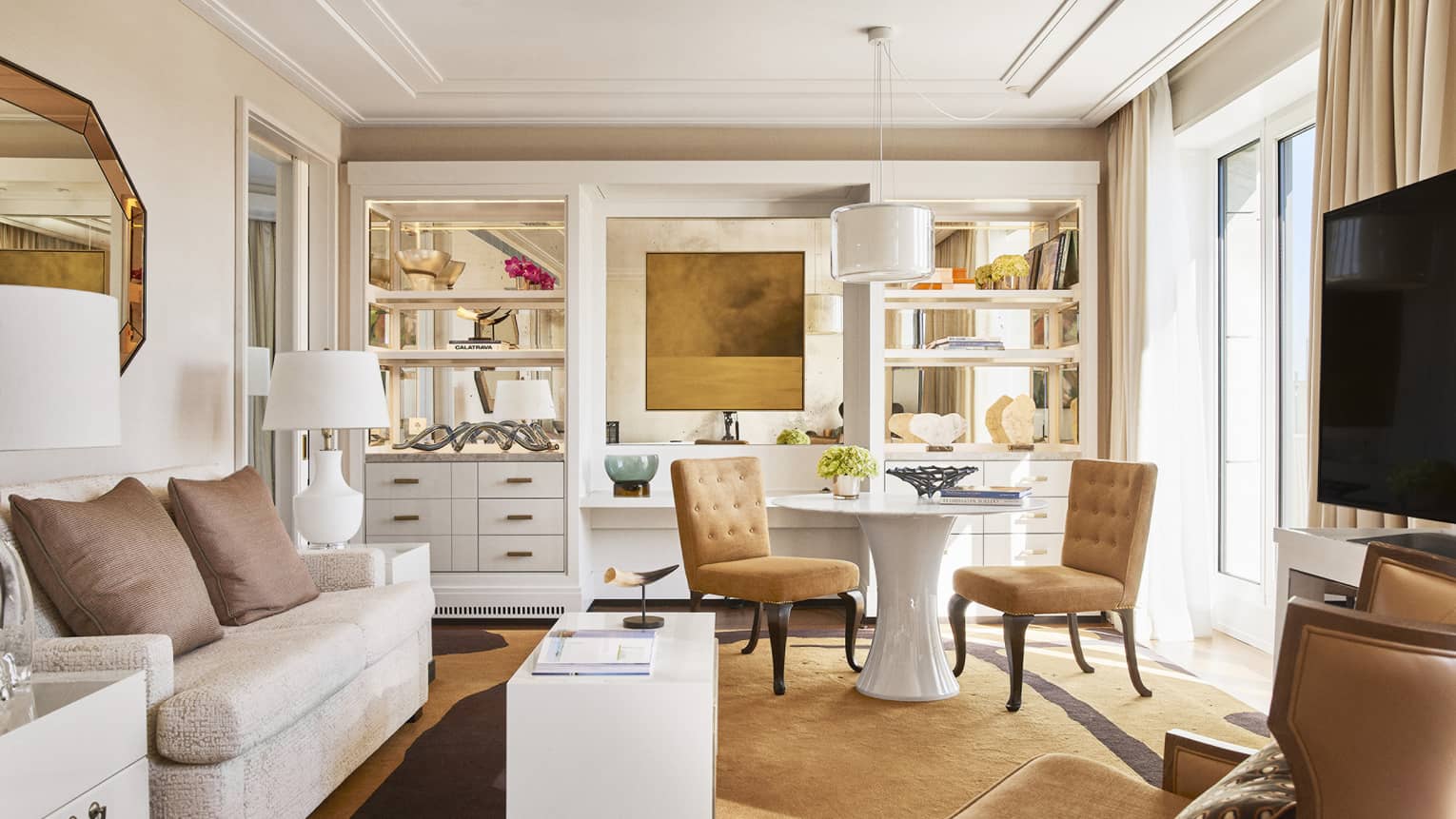 Luxury hotel living room with plush seating, tan chairs, round and rectangular tables, TV, decorative shelves and large windows with beige curtains
