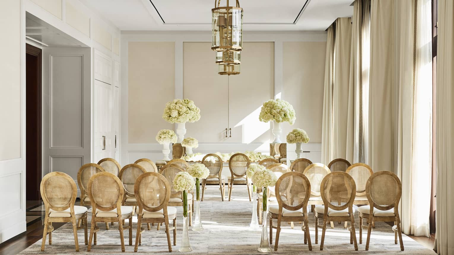 An elegant indoor wedding ceremony room with soft natural light, featuring wooden chairs arranged in rows, white floral arrangements and gold accents.