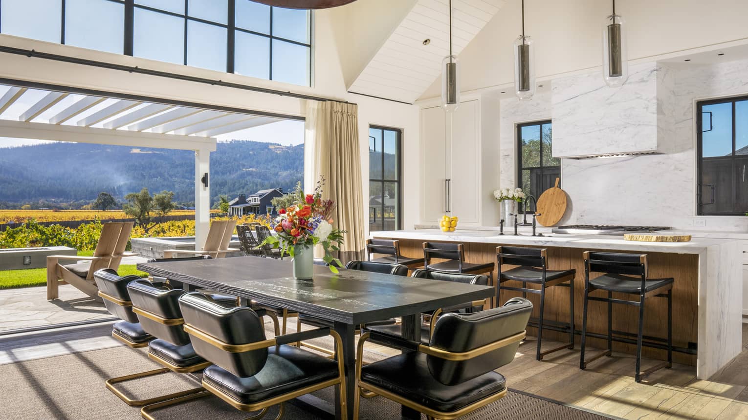 Kitchen and dining area with dark wood table, black leather chairs, marble island and open sliding doors leading to patio with vineyard and mountain views