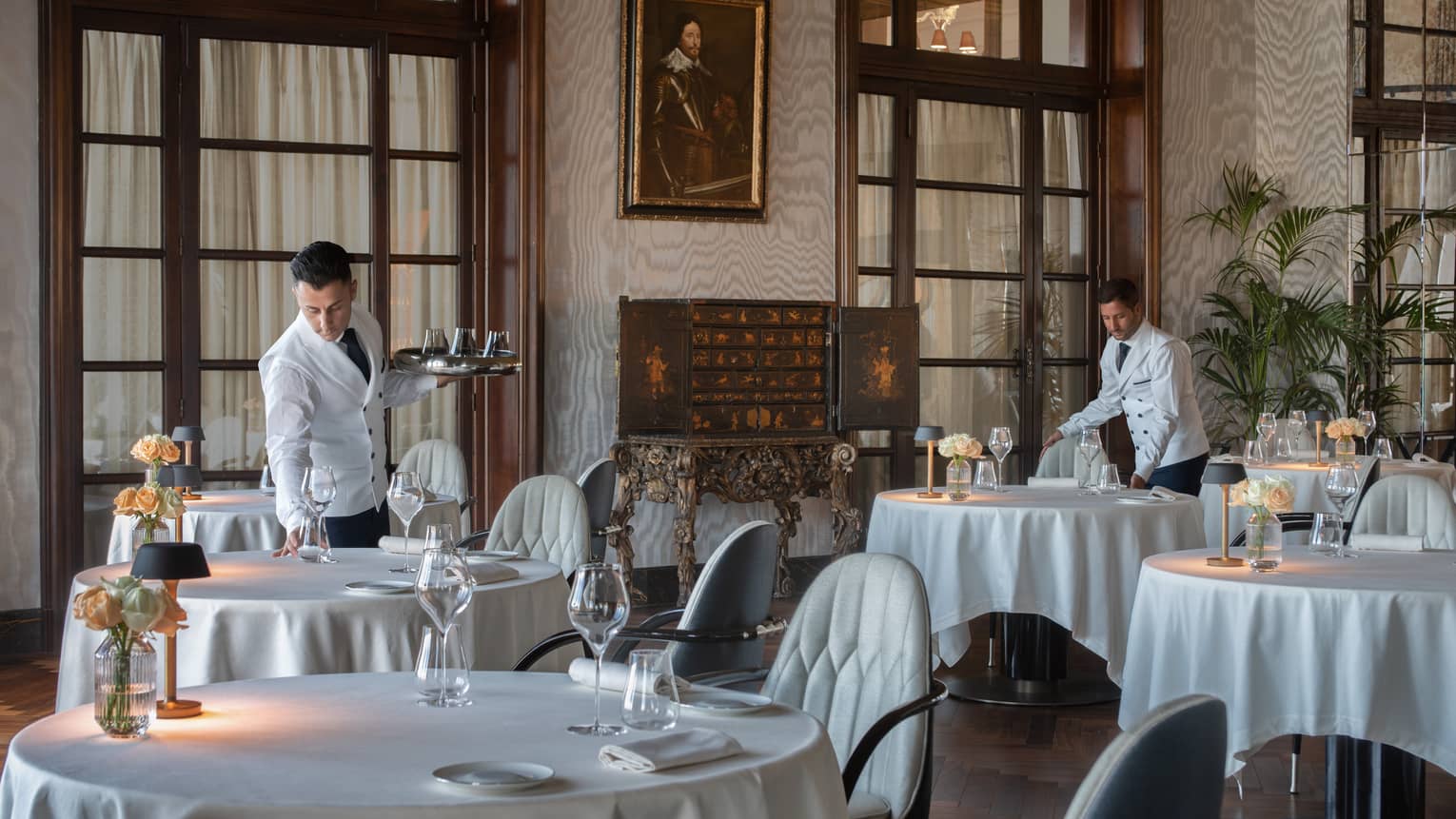 Two male servers in white jackets set dining tables in restaurant