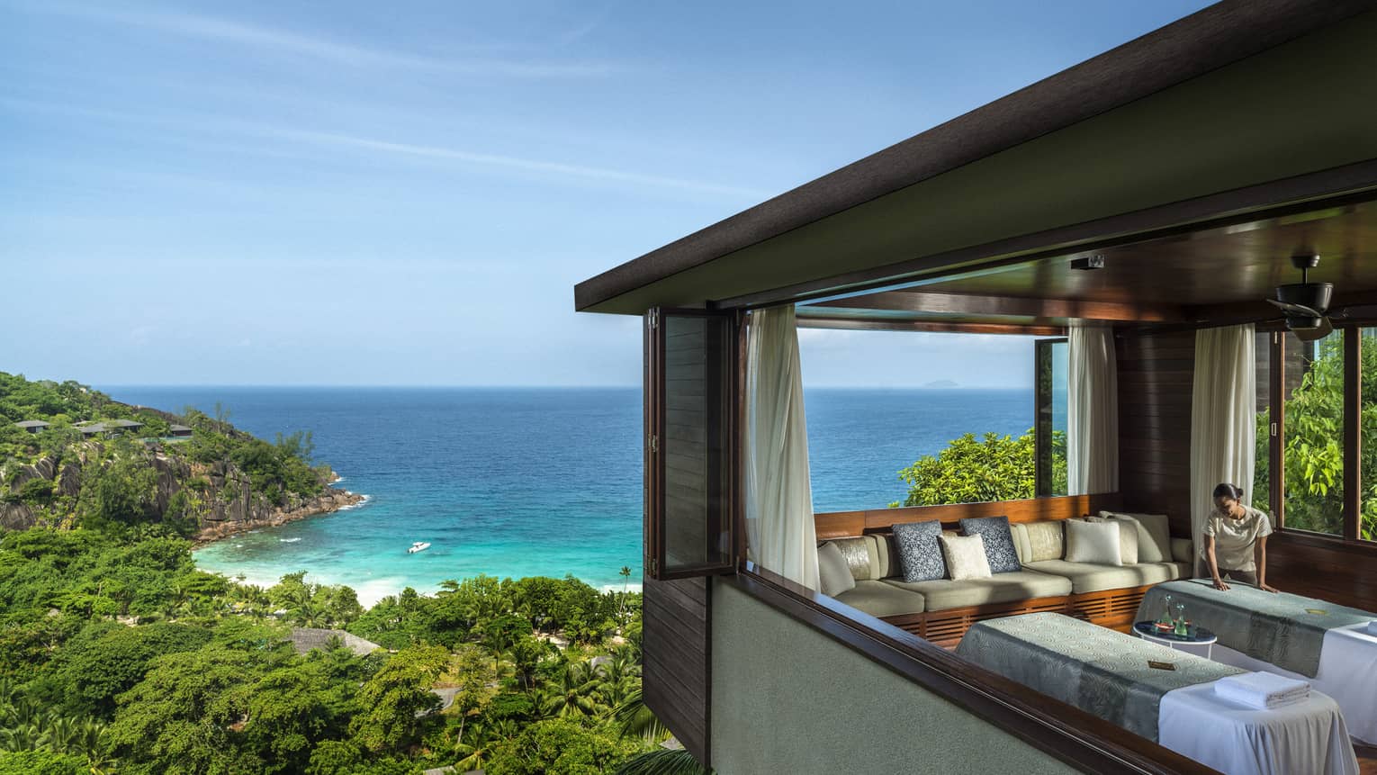Woman smoothes sheet on side-by-side massage tables in spa room high above trees, ocean