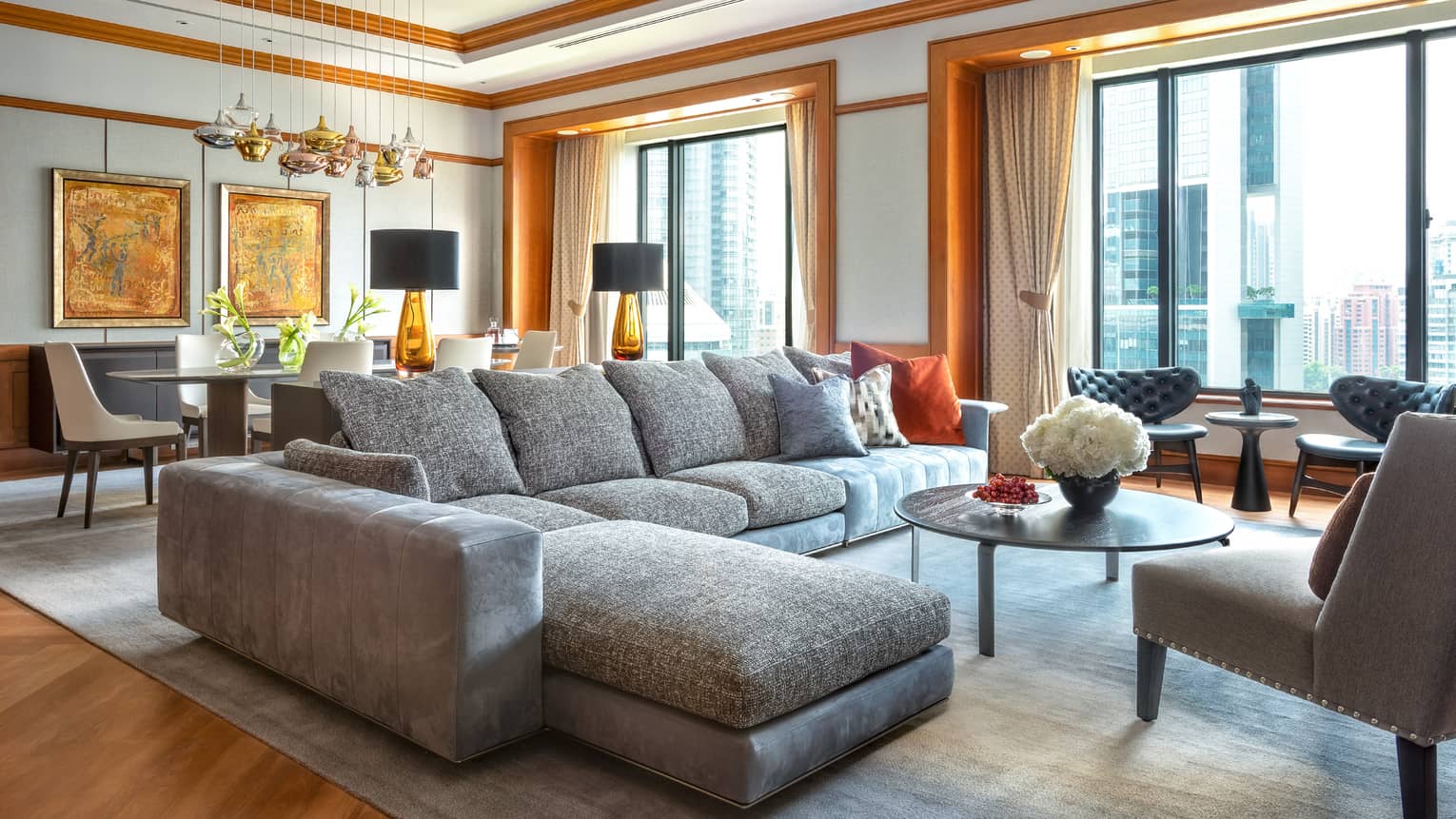A modern living room with a large grey sectional sofa, accented with colourful pillows, a round coffee table and floor-to-ceiling windows. A dining area with decorative lighting and artwork is visible in the background.
