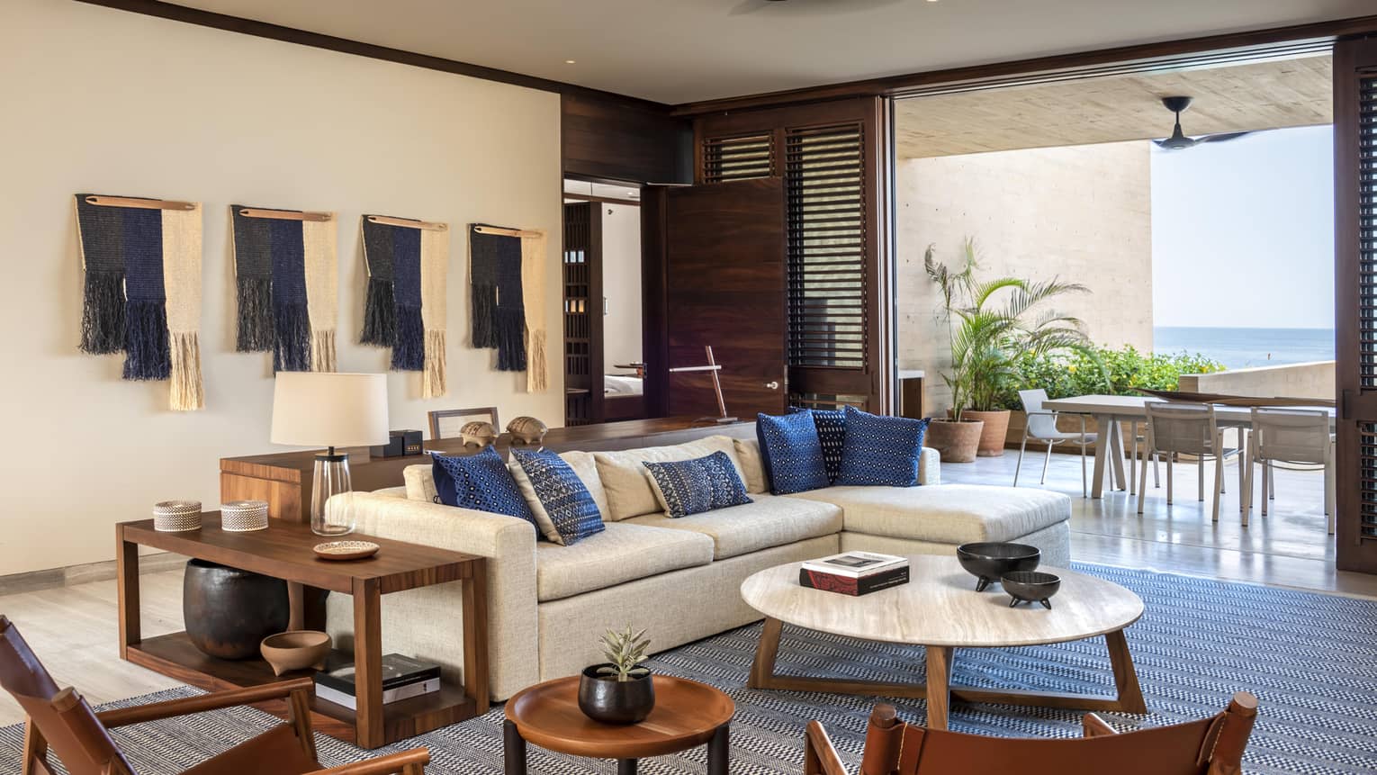 A modern living room with a beige sectional sofa decorated with blue accent pillows. Wooden furniture and wall hangings complement the space, which opens to a patio with ocean views.