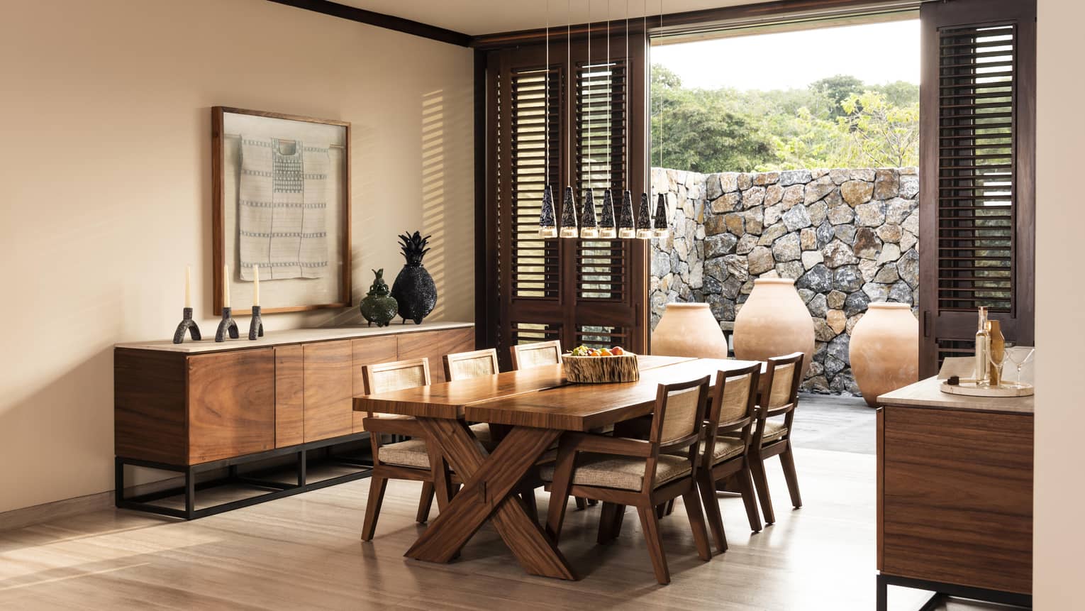 Elegant dining room with a wooden table, six chairs and a sideboard. Large vases and stone walls decorate the outdoor space, visible through wooden shutters. Hanging lights add warmth.