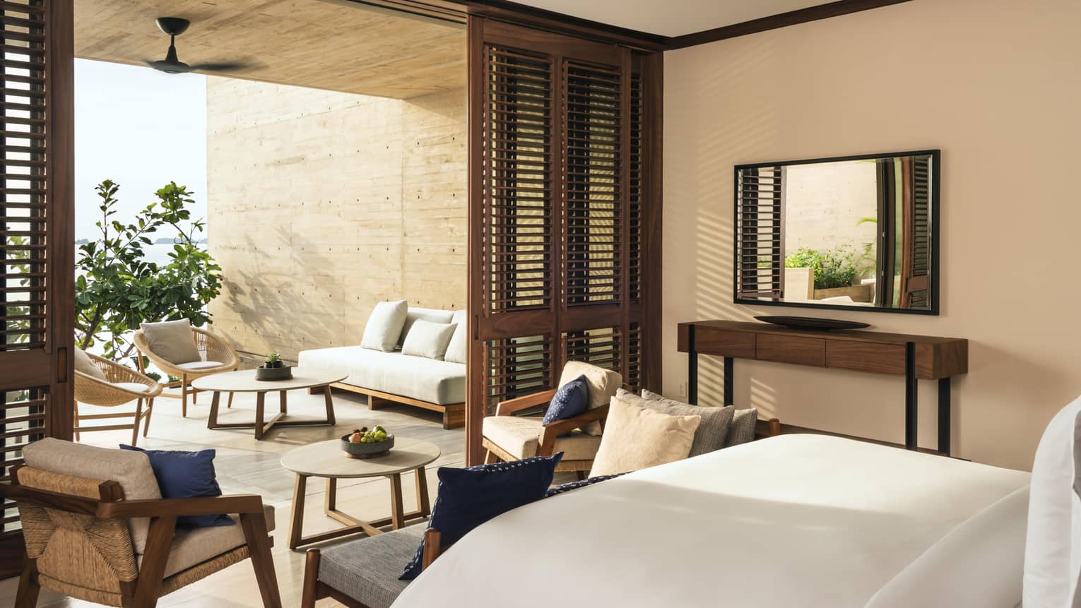 A spacious bedroom with wooden accents, opening to a terrace featuring wicker chairs, a sofa and round tables. The room has a modern, airy feel with soft natural light.