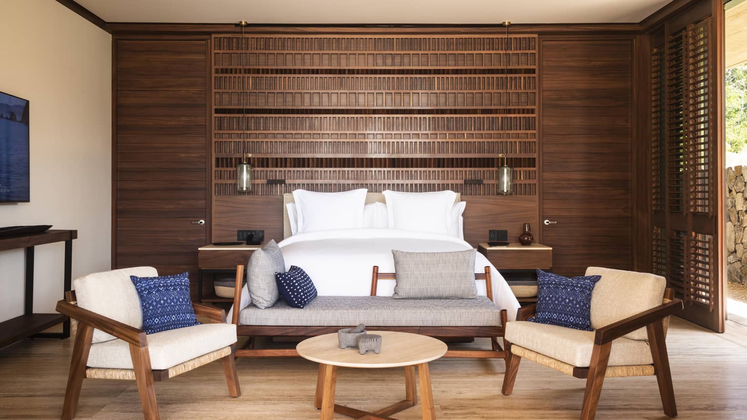 A modern bedroom with a wooden accent wall, featuring a large bed with white linens, two armchairs and a wooden coffee table. Blue decorative pillows add a pop of color.