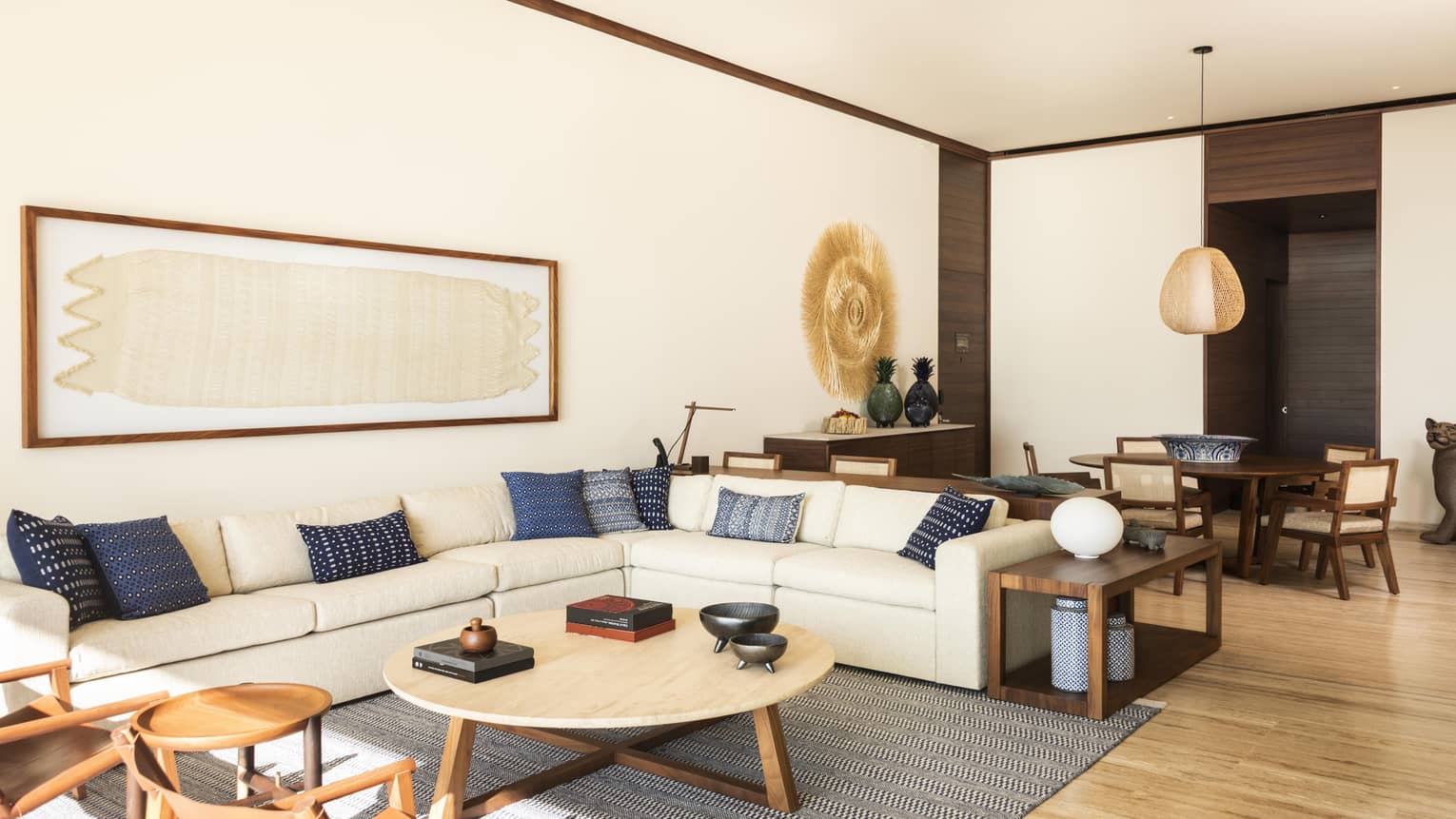 Bright living room with a beige sectional sofa, blue accent pillows and wooden furniture. A round coffee table sits on a patterned rug, and decorative wall art adds a natural touch.