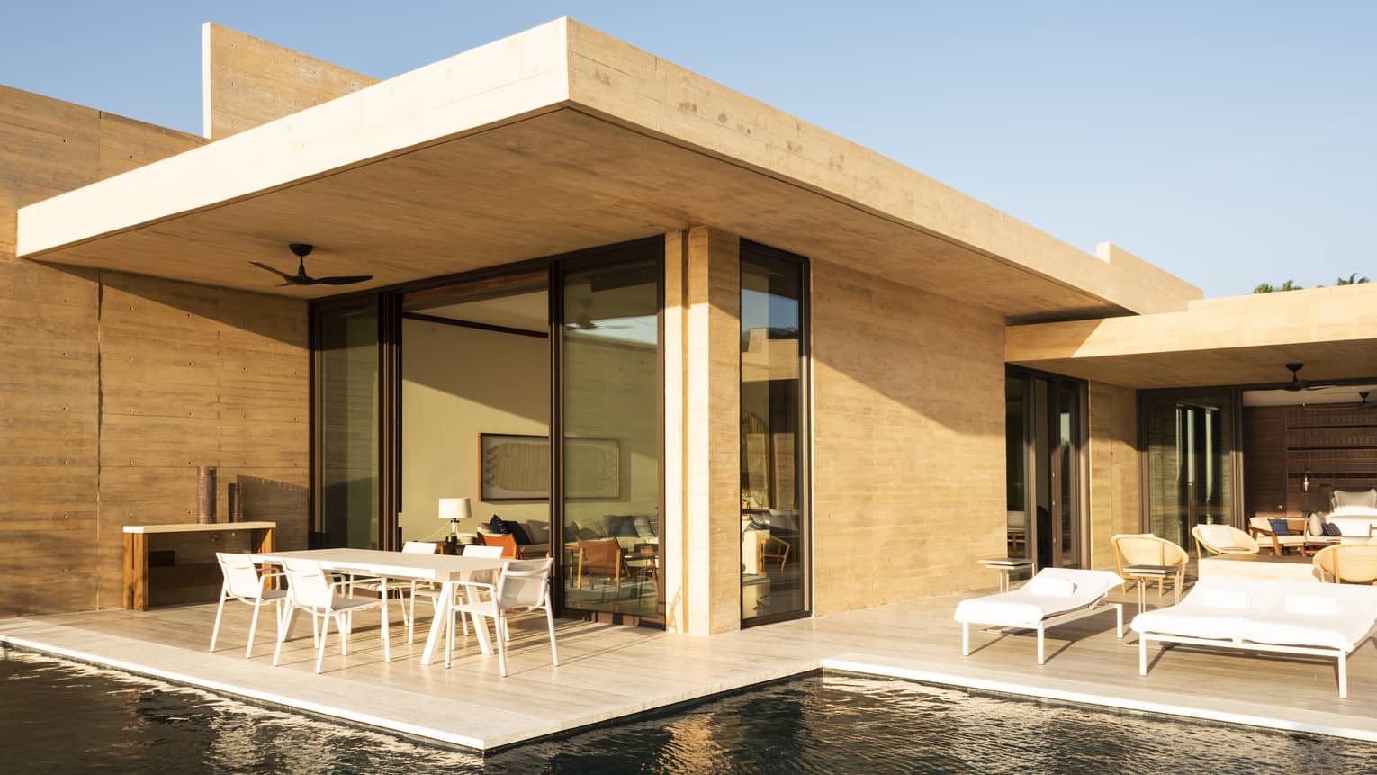Modern villa with a minimalist design, featuring an outdoor patio with white lounge chairs and a dining table, positioned next to a sleek pool under a clear sky