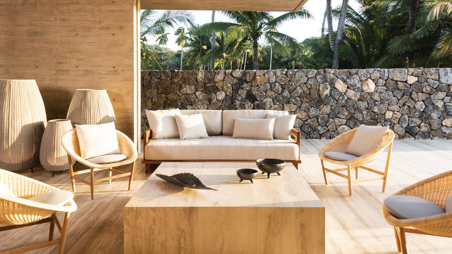 Outdoor seating area with a stone wall, light wooden furniture and wicker chairs around a stone coffee table. Large decorative woven lamps stand beside the sofa. Palm trees are visible in the background.