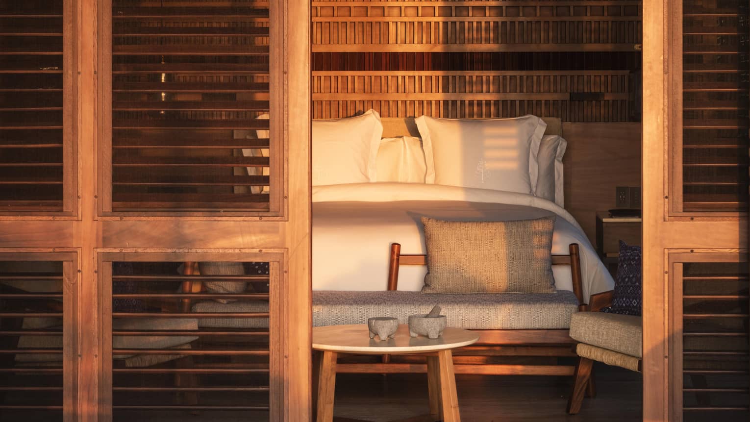 A cozy bedroom seen through wooden shutters, with warm sunlight casting shadows. The bed features white linens, and a wooden bench with pillows sits at the foot, accompanied by a small round table.