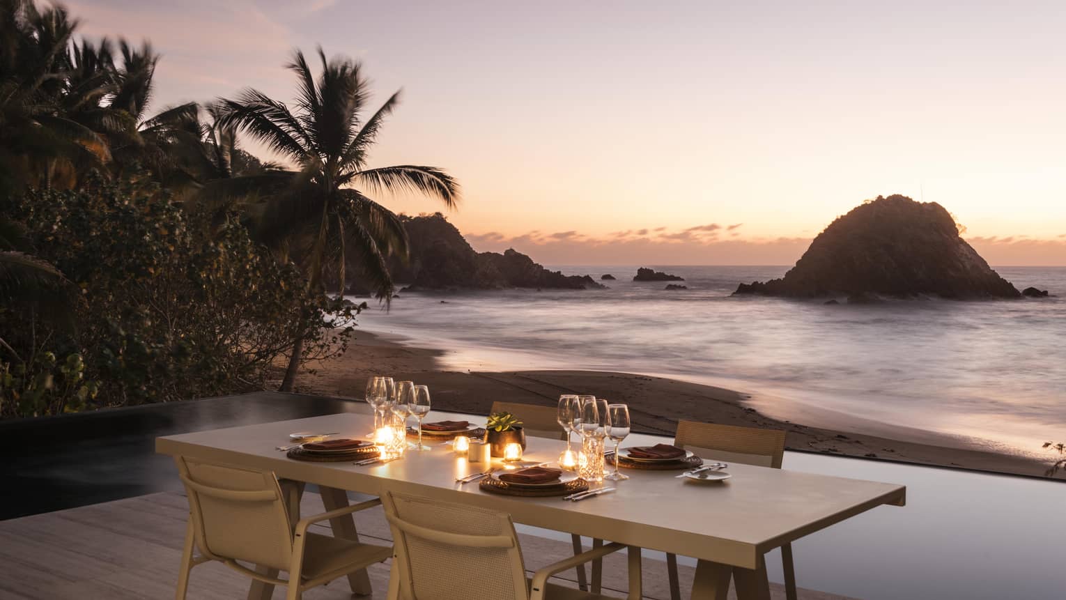 Outdoor dining table set with candles and glassware, overlooking a serene beach at sunset. Palm trees and rocky islets frame the shoreline, creating a tranquil ambience.