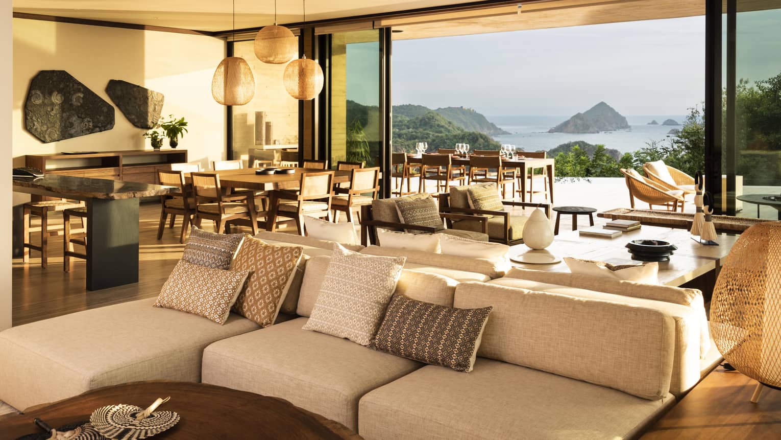 A spacious, open-concept living and dining area in a luxury villa, featuring cozy seating, wooden furnishings and large windows offering stunning views of the ocean and rocky islands