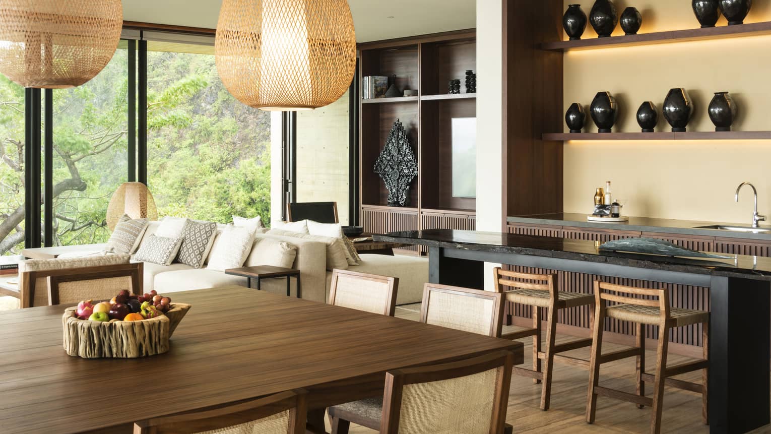 Stylish open-plan dining area with wooden furniture, wicker pendant lights and large windows offering views of lush greenery. A bowl of fresh fruit sits on the table.