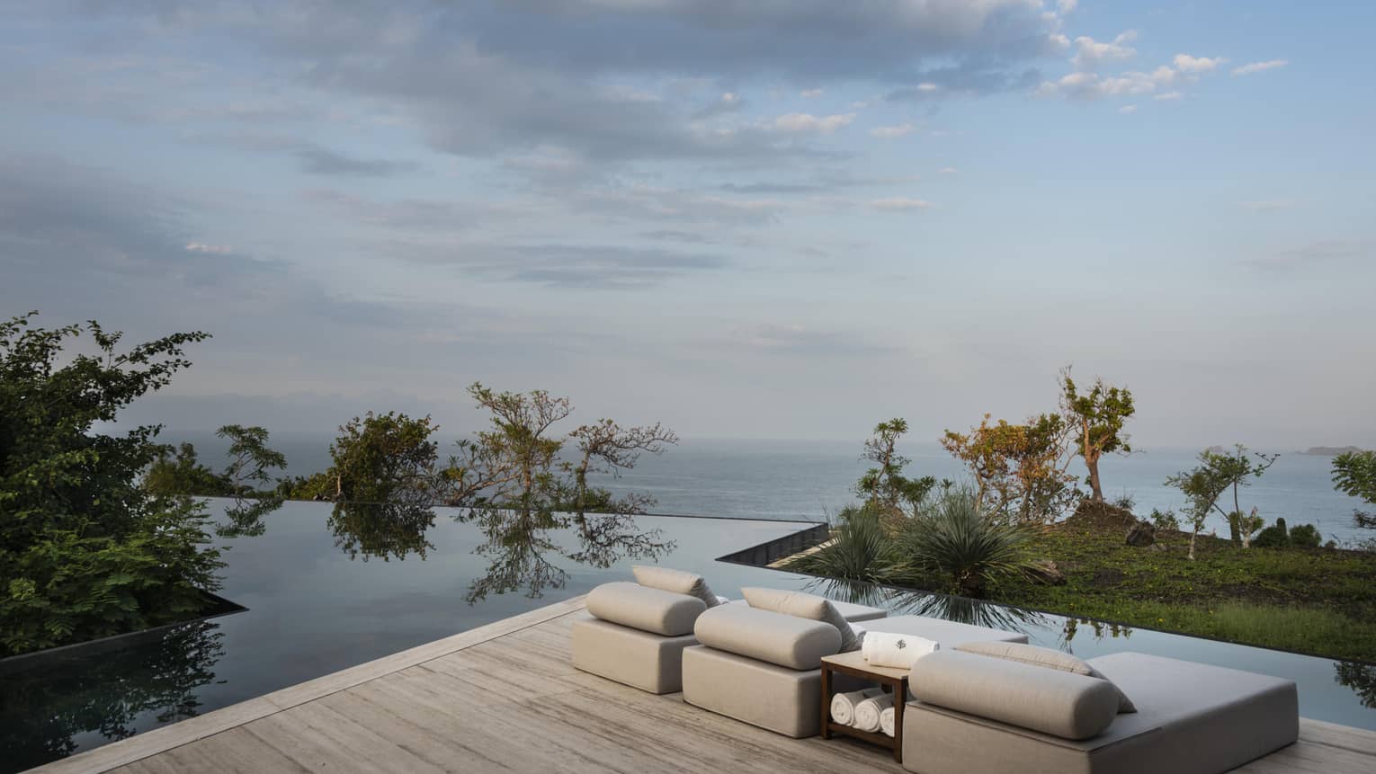 Minimalist poolside lounge area with cushioned seats overlooking an infinity pool and the ocean, surrounded by greenery and under a partially cloudy sky