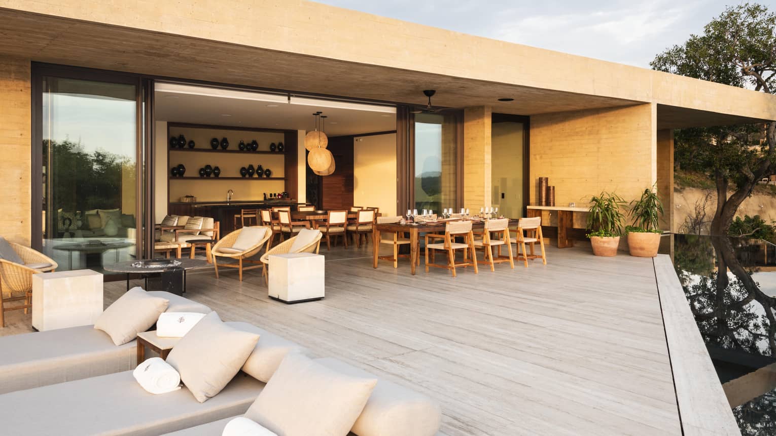 A luxurious outdoor terrace featuring cushioned lounge chairs, a dining area and wicker seating, seamlessly connecting to the indoor dining and kitchen space through large sliding doors