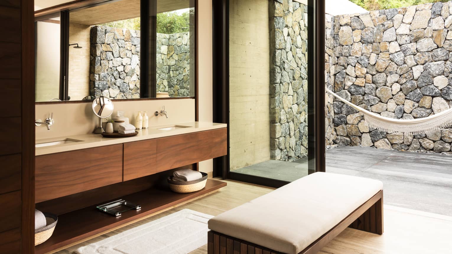Modern bathroom with double-sink vanity, wooden cabinets and a large mirror. Glass doors open to an outdoor patio with stone walls and a hammock.