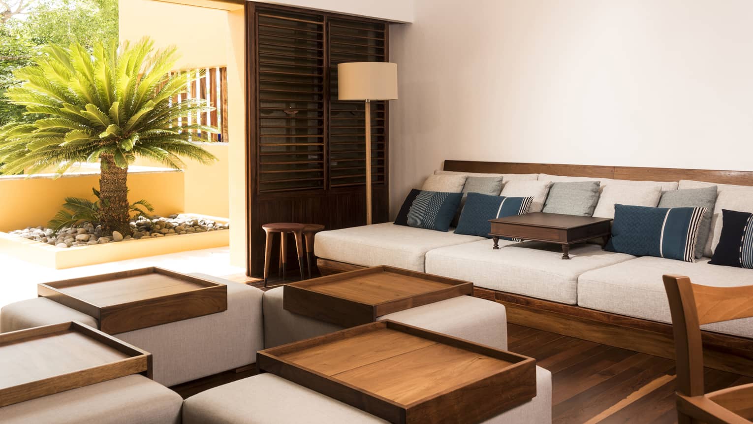 Living room with wooden sofa and ottomans, each with light grey cushions, and walk-out terrace