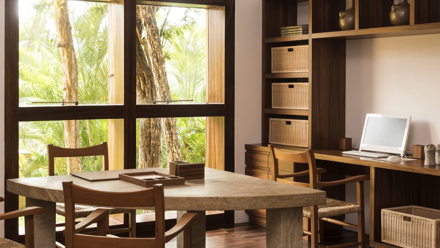 Room with a wooden desk, wicker basket organization, table and two chairs, windows looking out to jungle forest