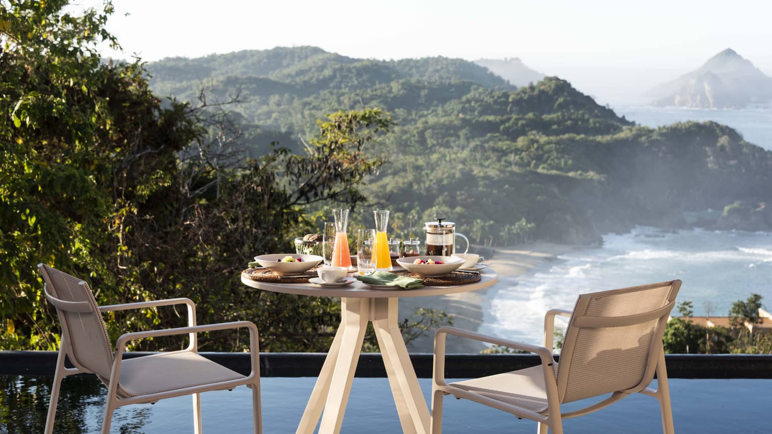 A small table with drinks and food on it looking out a beach and trees.