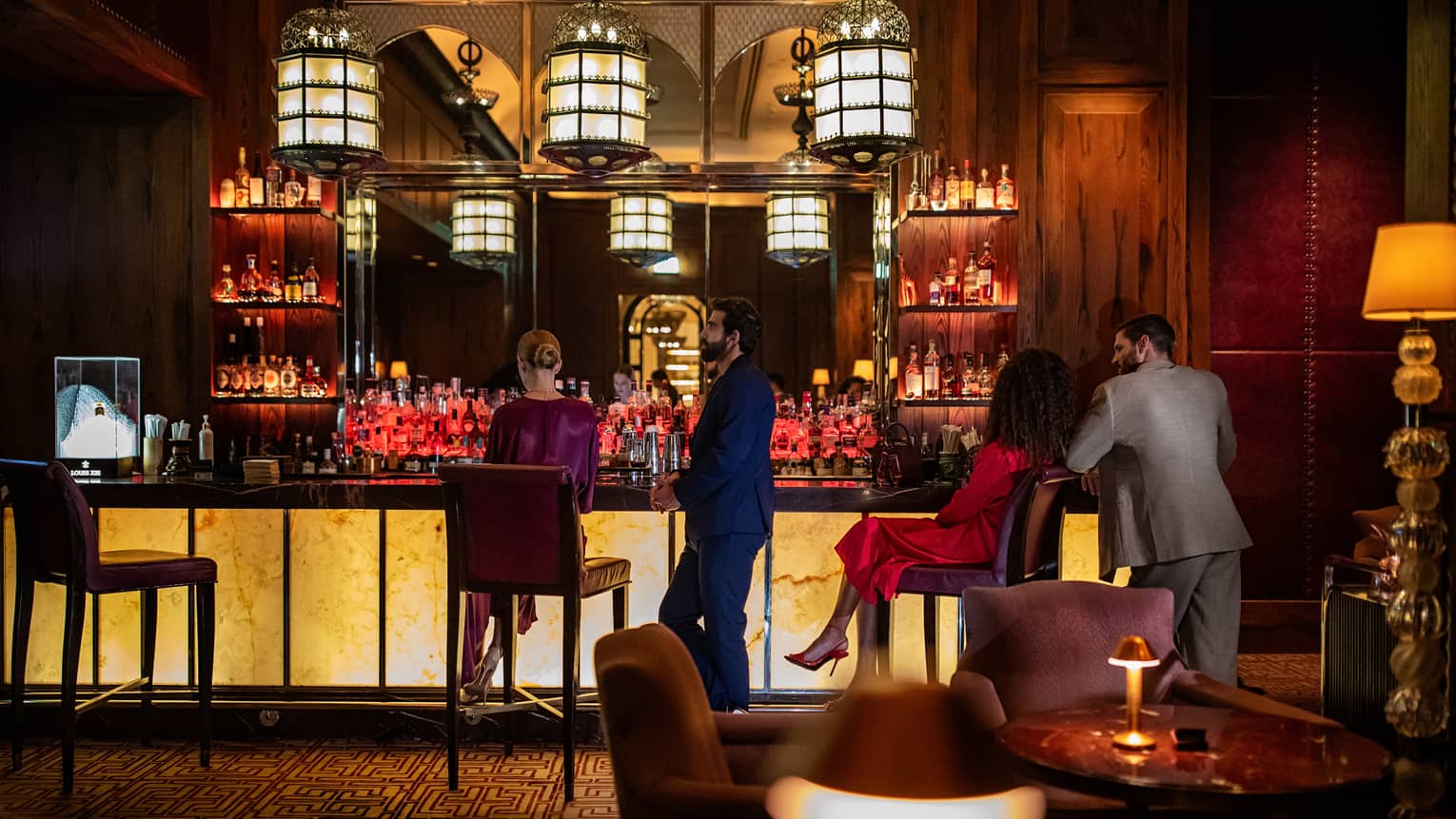 Two men and two women sit at an illuminated bar in front of several shelves of bottles.