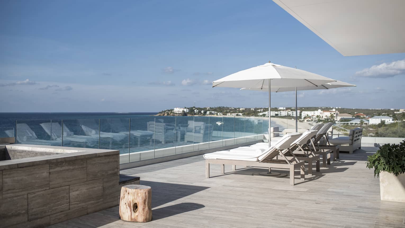 Private sea-view terrace with lounge chairs, a white umbrella and glass railings overlooking the ocean and a coastal landscape