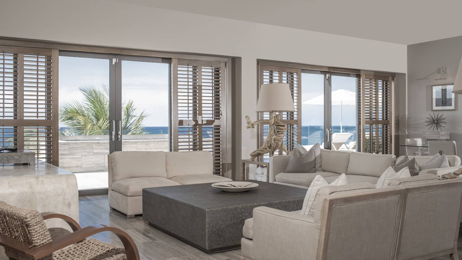 Luxury living room with white sofas, large square coffee table, large windows with wooden shutters and a view of the ocean and patio with lounge chairs