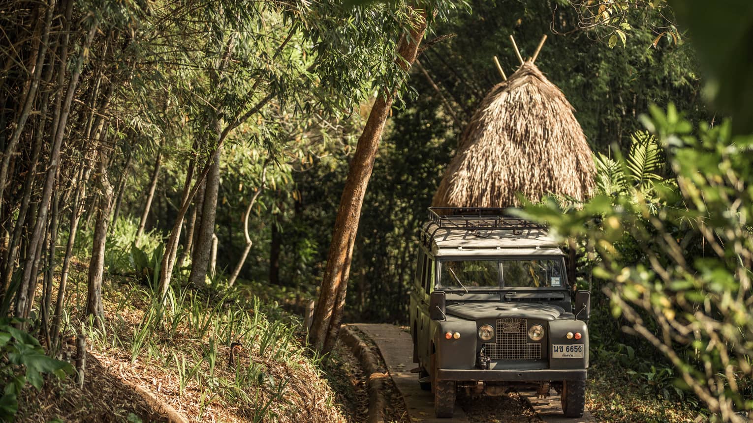 Jeep driving down path in forest past thatched-roof hut