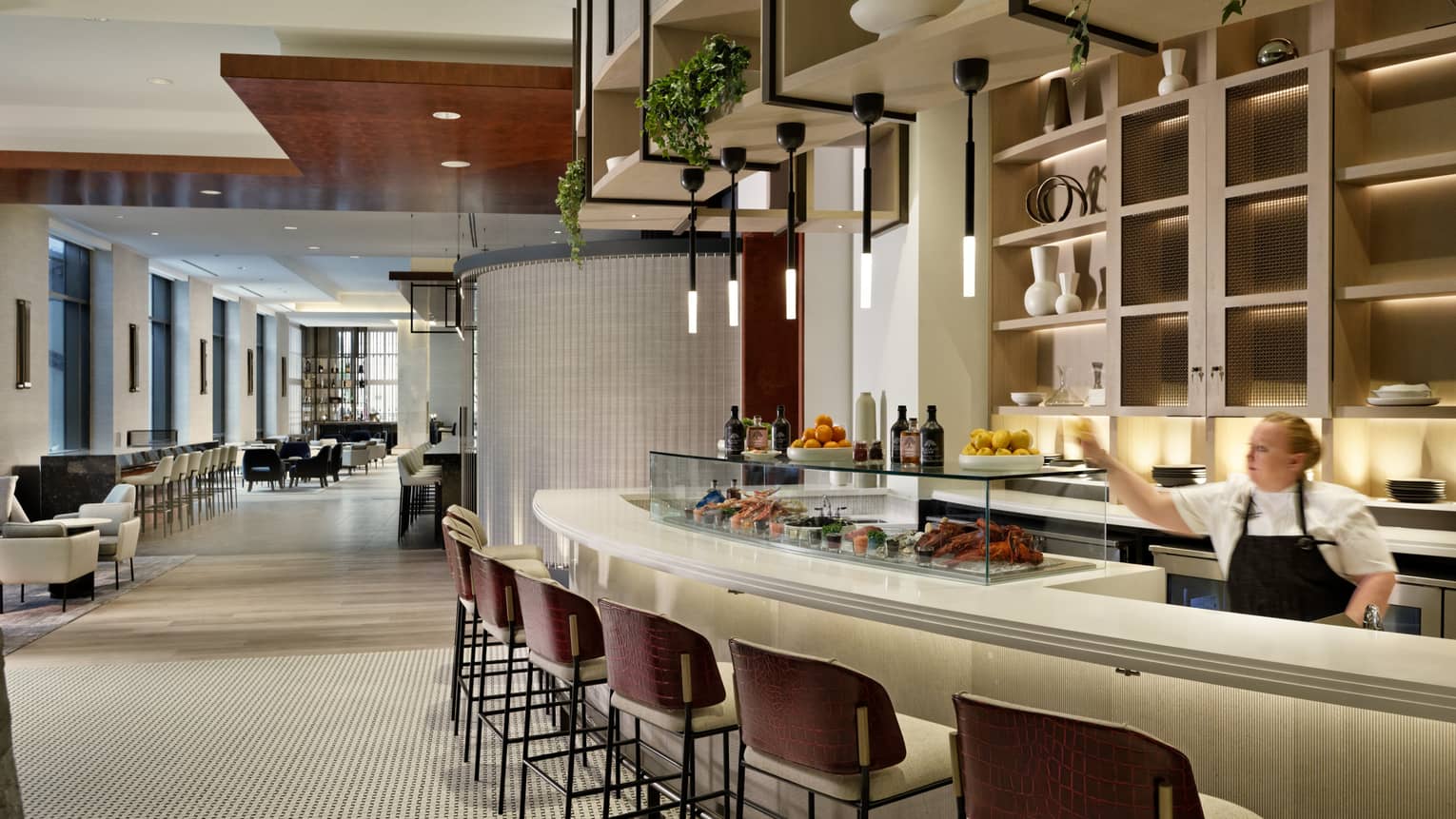 A staff member working behind a curved bar with bar stools, food is displayed on the counter.